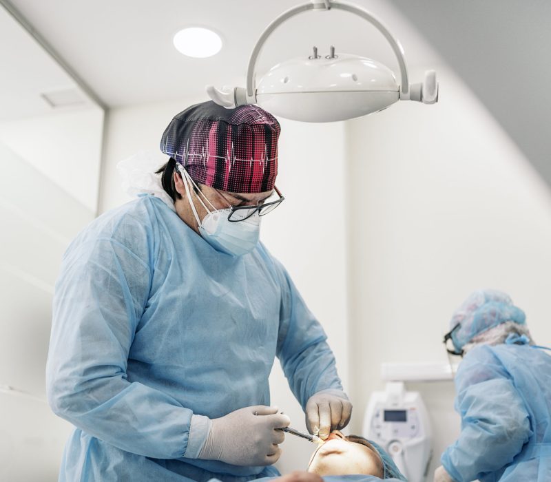 Male dentist and his team wearing face mask and protective clothes doing treatment to unrecognized patient.